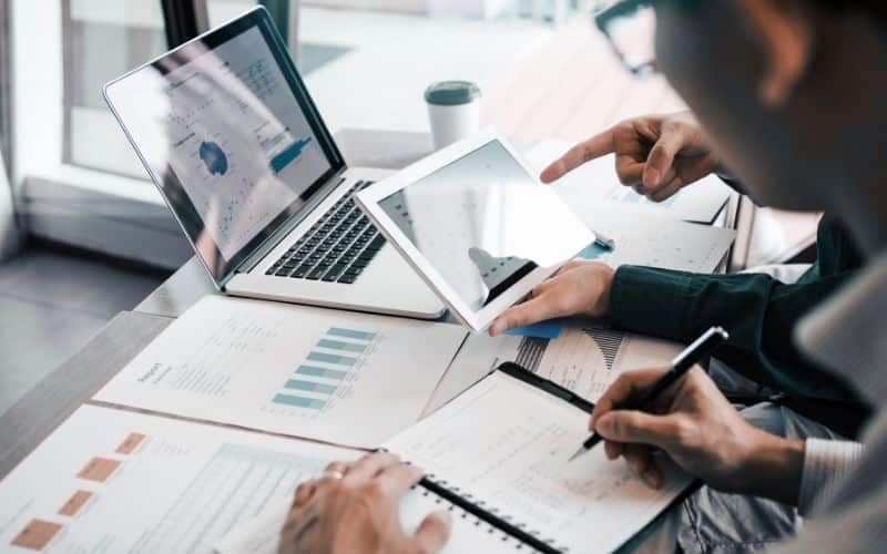 two workers look at an ipad and data sheets to conduct a cost comparison for outsourcing packing to a contract manufacturer.