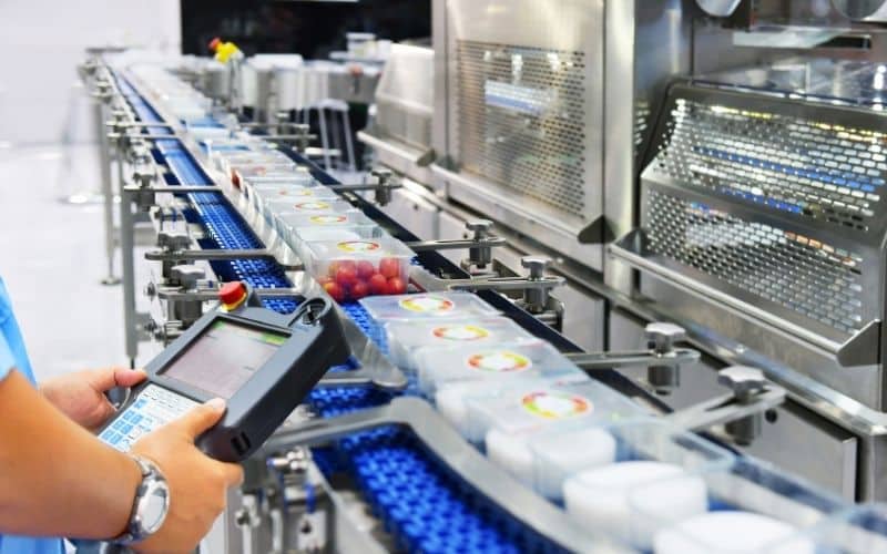 A contract manufacturing facility conducts a routine quality test. An employee holds a tablet while overseeing food packaging on a conveyor belt.