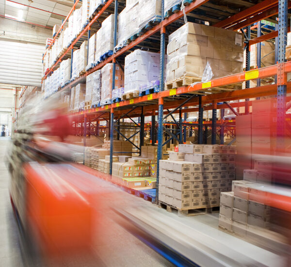 A warehouse with tall shelves stacked with boxes and pallets. A red forklift, essential for distribution, moves quickly in the foreground, appearing blurred due to motion, while the background shows organized inventory.