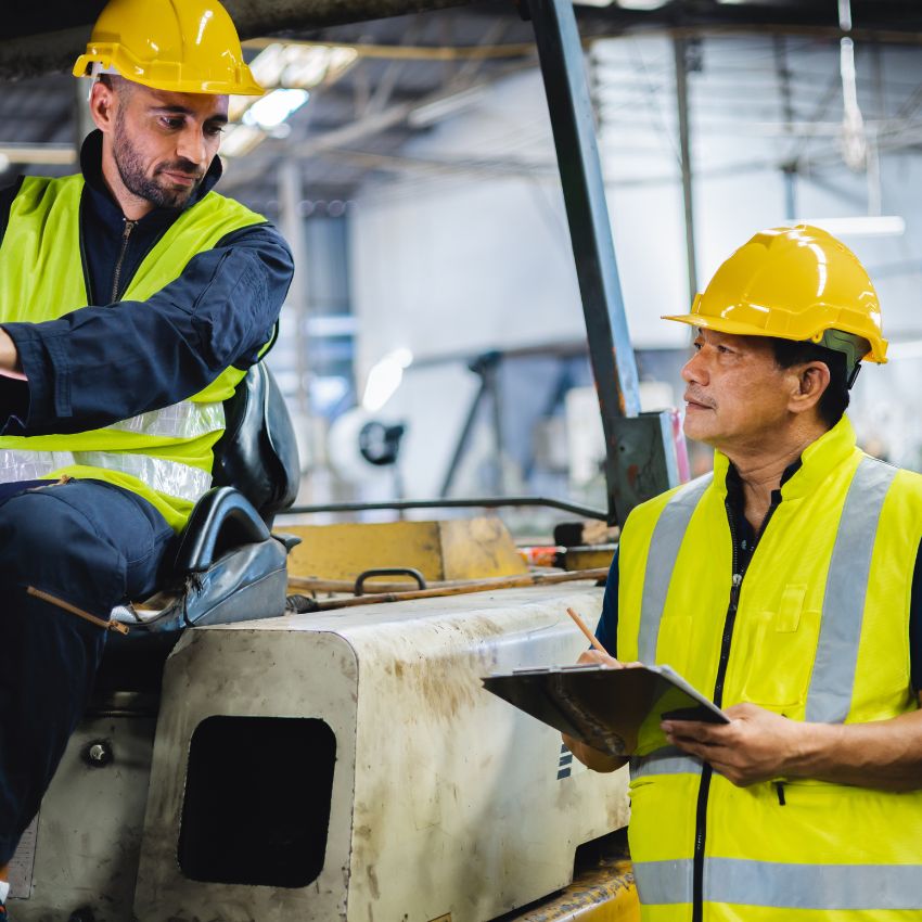 Two construction workers wearing yellow safety vests and hard hats are having a discussion about contract manufacturing. One is seated on industrial machinery, while the other stands nearby with a clipboard in an indoor industrial setting.