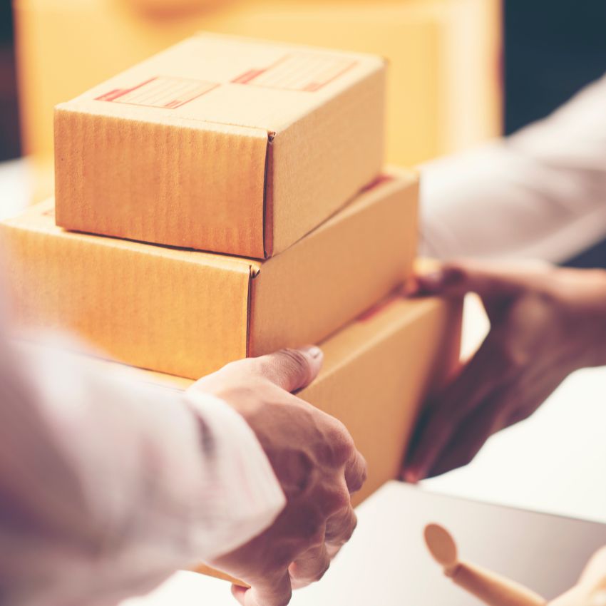 Two people exchanging a stack of three cardboard boxes, illustrating custom processing as one pair of hands gives and another receives packages in a well-lit indoor setting.