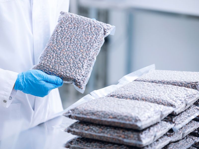 A food manufacturing employee wearing blue gloves holds a vacuum sealed bag of coffee beans.