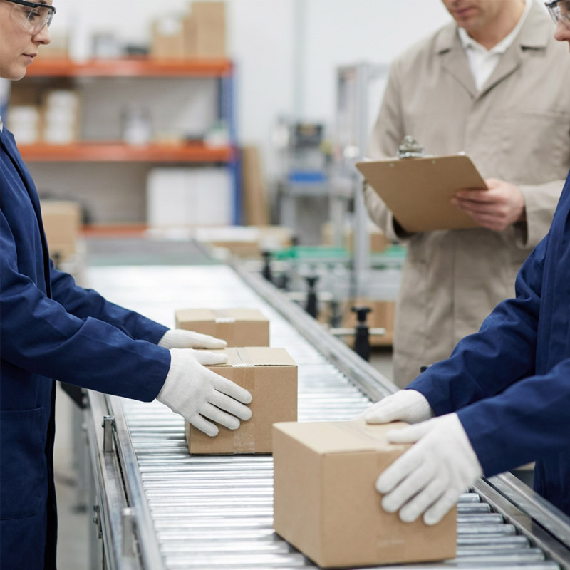 Workers in blue uniforms and white gloves handle cardboard boxes on a conveyor belt while another person in a beige coat with a clipboard oversees the turnkey manufacturing process in a warehouse or factory setting.