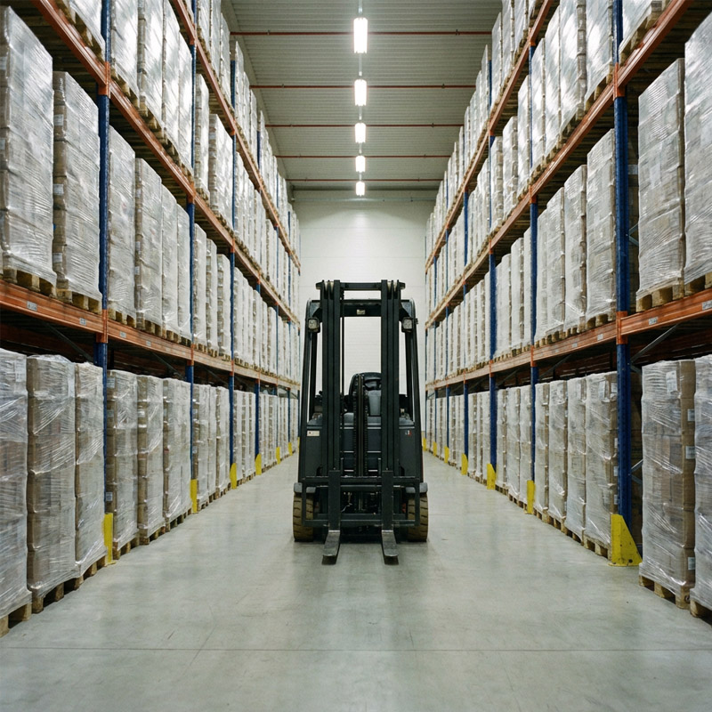 A forklift is parked in the center aisle of a large warehouse, surrounded by tall shelves stacked with palletized goods wrapped in plastic. The warehouse is brightly lit with overhead lights.