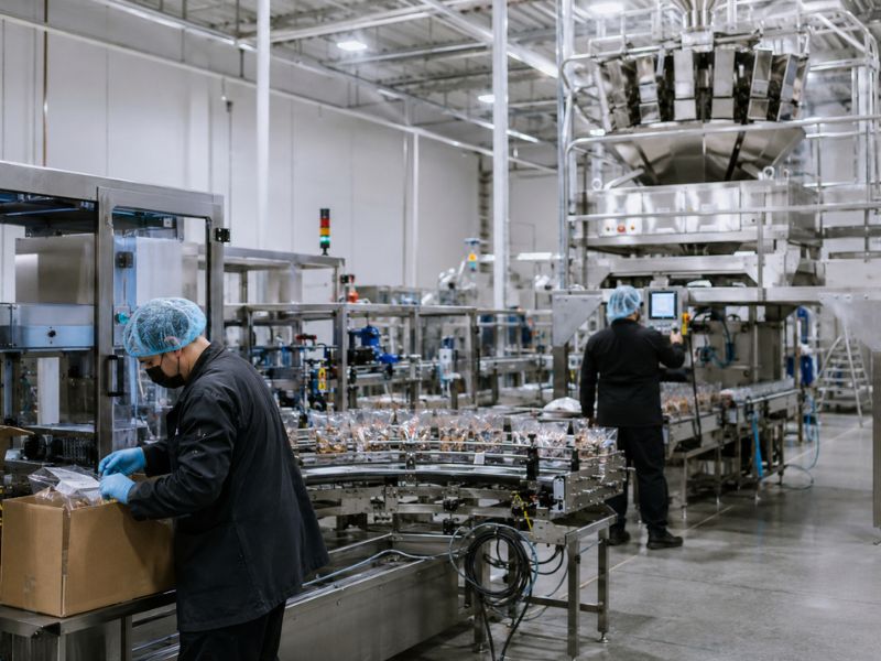 Two factory workers in protective gear operate machinery and package products on a modern food processing assembly line inside a large, brightly lit industrial facility, exemplifying efficient Contract Packaging services.